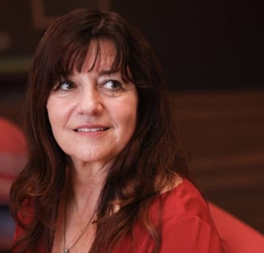 21b Consulting - An image of Sandee Bowman - A woman with long, dark brown hair and bangs is looking off to the side. She is wearing a red top and a necklace. The background is blurred, with hints of red and dark colors.