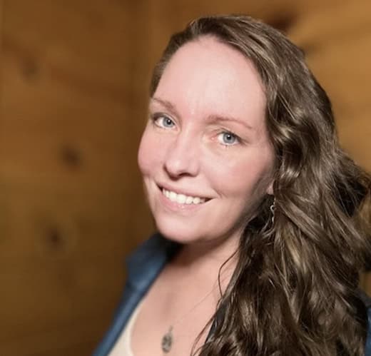 21b Consulting - An image of Noelle Woodhead - A woman with long wavy brown hair and blue eyes is smiling at the camera. She is wearing a blue top and a necklace, and the background appears to be a wooden room with a softly blurred effect.