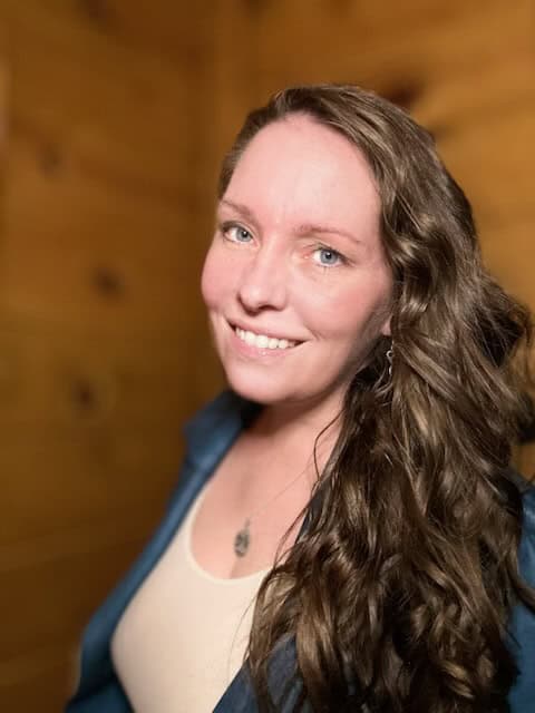 21b Consulting – A person with long, wavy brown hair is smiling at the camera. They are wearing a light-colored top and a blue jacket, with a necklace featuring a small pendant. The background is wooden, possibly indicating an indoor setting. 21b Consulting - A person with long, wavy brown hair is smiling at the camera. They are wearing a light-colored top and a blue jacket, with a necklace featuring a small pendant. The background is wooden, possibly indicating an indoor setting.