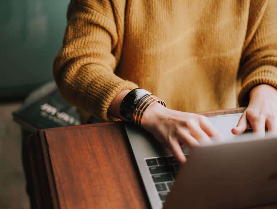 21b Consulting – A person in a mustard sweater types on a laptop, with circles of bracelets adorning their left wrist. Nearby, an Info Element book rests on the wooden table. The setting exudes a cozy, productive atmosphere. 21b Consulting - A person in a mustard sweater types on a laptop, with circles of bracelets adorning their left wrist. Nearby, an Info Element book rests on the wooden table. The setting exudes a cozy, productive atmosphere.